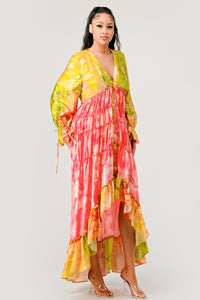 Sunset Breeze Tiered Maxi Dress | Model wearing a vibrant bohemian-style maxi dress with a colorful tie-dye pattern in shades of yellow, pink, and green. Featuring a deep V-neckline, tiered ruffle detailing, and a high-low hem, this dress exudes a carefree and whimsical vibe, perfect for summer events or festivals. Rêve Dorés