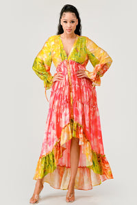 Sunset Breeze Tiered Maxi Dress | Model wearing a vibrant bohemian-style maxi dress with a colorful tie-dye pattern in shades of yellow, pink, and green. Featuring a deep V-neckline, tiered ruffle detailing, and a high-low hem, this dress exudes a carefree and whimsical vibe, perfect for summer events or festivals. Rêve Dorés