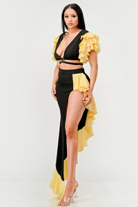 Sunset Ruffle Glamour Gown - Model wearing a sophisticated black and yellow evening gown featuring a plunging neckline with a silver ring accent, a high side slit, and ruffled yellow details cascading along the shoulders and hem. Perfect for elegant summer events or formal occasions. Rêve Dorés