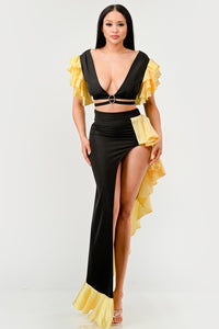 Sunset Ruffle Glamour Gown - Model wearing a sophisticated black and yellow evening gown featuring a plunging neckline with a silver ring accent, a high side slit, and ruffled yellow details cascading along the shoulders and hem. Perfect for elegant summer events or formal occasions. Rêve Dorés