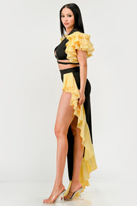 Sunset Ruffle Glamour Gown - Model wearing a sophisticated black and yellow evening gown featuring a plunging neckline with a silver ring accent, a high side slit, and ruffled yellow details cascading along the shoulders and hem. Perfect for elegant summer events or formal occasions. Rêve Dorés