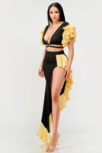 Sunset Ruffle Glamour Gown - Model wearing a sophisticated black and yellow evening gown featuring a plunging neckline with a silver ring accent, a high side slit, and ruffled yellow details cascading along the shoulders and hem. Perfect for elegant summer events or formal occasions. Rêve Dorés