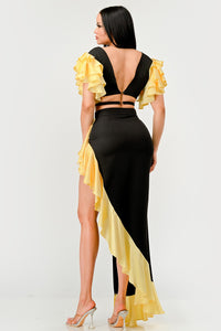 Sunset Ruffle Glamour Gown - Model wearing a sophisticated black and yellow evening gown featuring a plunging neckline with a silver ring accent, a high side slit, and ruffled yellow details cascading along the shoulders and hem. Perfect for elegant summer events or formal occasions. Rêve Dorés