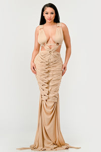 Sahara Mystique Pleated Gown - Model wearing a stunning beige cutout maxi dress featuring a plunging neckline, circular ring accents, and intricate ruched detailing throughout. The floor-length design with a figure-hugging silhouette exudes elegance and bold sophistication, perfect for an upscale evening event. - Rêves Dorés