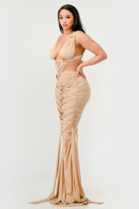 Sahara Mystique Pleated Gown - Model wearing a stunning beige cutout maxi dress featuring a plunging neckline, circular ring accents, and intricate ruched detailing throughout. The floor-length design with a figure-hugging silhouette exudes elegance and bold sophistication, perfect for an upscale evening event. - Rêves Dorés