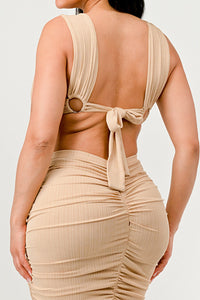 Sahara Mystique Pleated Gown - Model wearing a stunning beige cutout maxi dress featuring a plunging neckline, circular ring accents, and intricate ruched detailing throughout. The floor-length design with a figure-hugging silhouette exudes elegance and bold sophistication, perfect for an upscale evening event. - Rêves Dorés