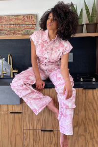 Pink Whisper Tie-Dye Jumpsuit | Model wearing a pink utility-style jumpsuit featuring a button-up front, short sleeves, and cargo pockets for a functional yet stylish look. The relaxed fit and soft tie-dye pattern make it perfect for casual outings or chic everyday wear by Rêve Dorés