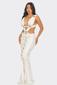 Sahara Mystique Pleated Gown - Model wearing a stunning beige cutout maxi dress featuring a plunging neckline, circular ring accents, and intricate ruched detailing throughout. The floor-length design with a figure-hugging silhouette exudes elegance and bold sophistication, perfect for an upscale evening event. - Rêves Dorés