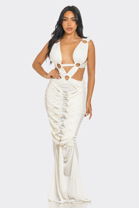 Sahara Mystique Pleated Gown - Model wearing a stunning beige cutout maxi dress featuring a plunging neckline, circular ring accents, and intricate ruched detailing throughout. The floor-length design with a figure-hugging silhouette exudes elegance and bold sophistication, perfect for an upscale evening event. - Rêves Dorés