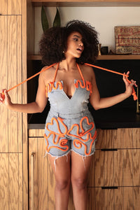 Rêve Dorés model wearing a unique denim halter romper featuring a deep V-neckline, frayed edges, and bold orange rope detailing styled in abstract patterns across the bodice and shorts. The playful design combines edgy and artistic elements, perfect for making a bold fashion statement. 