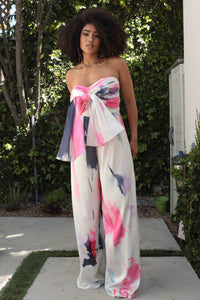 Wide Leg Abstract Elegance Printed Jumpsuit. Model wearing a strapless wide-leg jumpsuit featuring a bold abstract print in pink, white, and black. The design includes a front tie detail at the bust, creating an elegant and artistic look, perfect for summer events or resort wear by Rêve Dorés