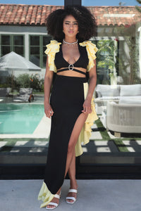 Model wearing a sophisticated black and yellow evening gown featuring a plunging neckline with a silver ring accent, a high side slit, and ruffled yellow details cascading along the shoulders and hem. Perfect for elegant summer events or formal occasions. Rêve Dorés